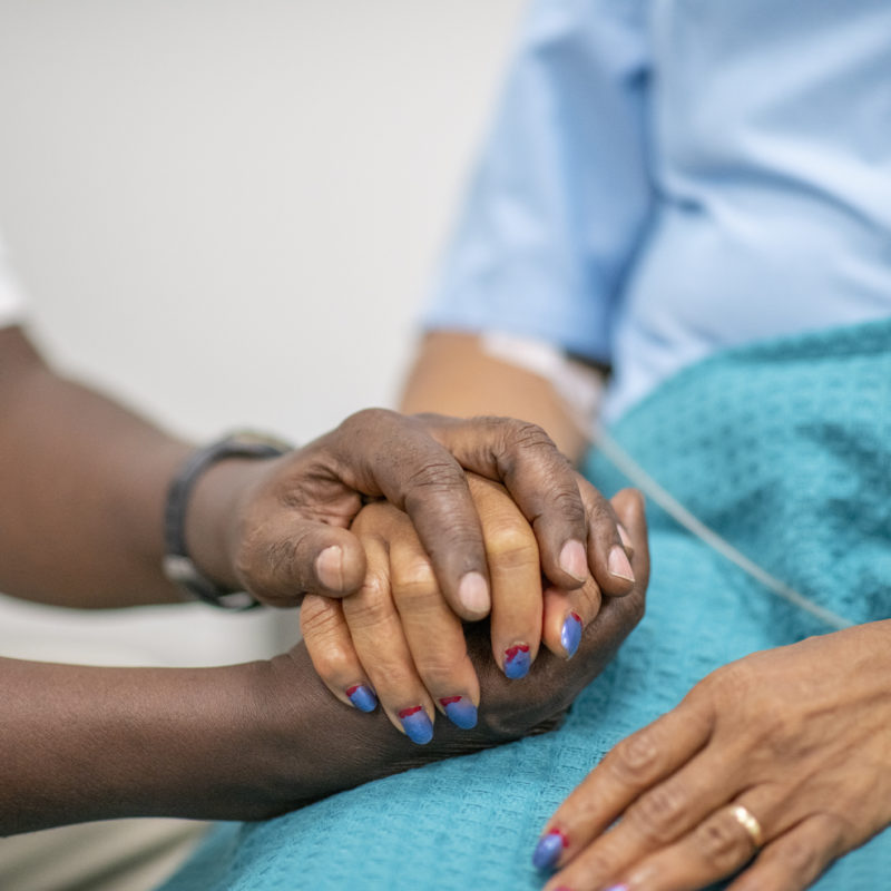 Elderly woman is comforted by a medical professional during the Covid-19. Focus is on their hands. The medical staff is holding the woman